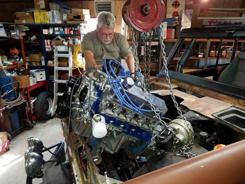 1972 Ford Bronco - A mechanic is installing a new engine into a vehicle using a chain hoist in a workshop.