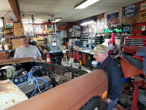 1972 Ford Bronco - A cluttered garage with tools and Ford memorabilia on the walls, as two men attend to a vehicle's engine.