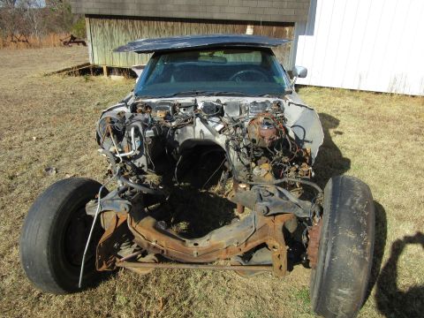 Car chassis with front clip removed, showing engine bay and suspension components. Hood is bent upwards