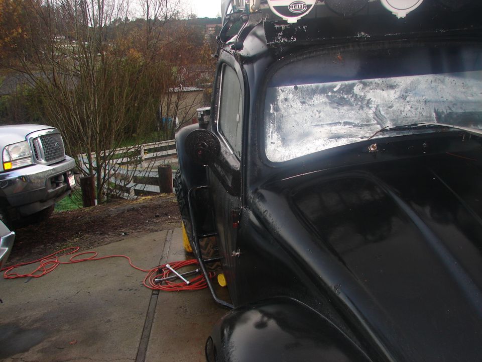 Black vintage car with roof rack and Hella lights, parked next to a silver truck