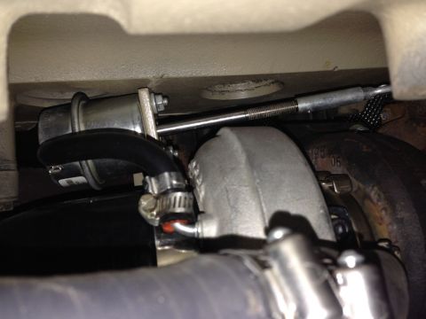 1994 Ford Bronco - A close-up view of a turbocharger wastegate actuator rod connected to a turbocharger housing. A black rubber hose with a clamp is also visi…
