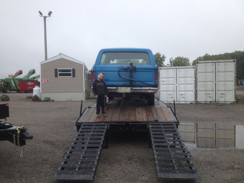 Boy standing on a trailer next to a blue Ford Bronco.
