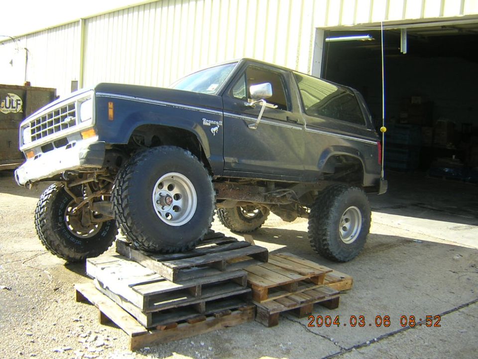 Dark blue Ford Bronco II with lifted suspension and large tires, parked on wooden pallets in front of a garage