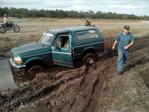 1996 Ford Bronco - A teal 1996 Ford Bronco is stuck in deep mud with its driver's side door open. A man in a blue t-shirt walks away from the vehicle on a mud…