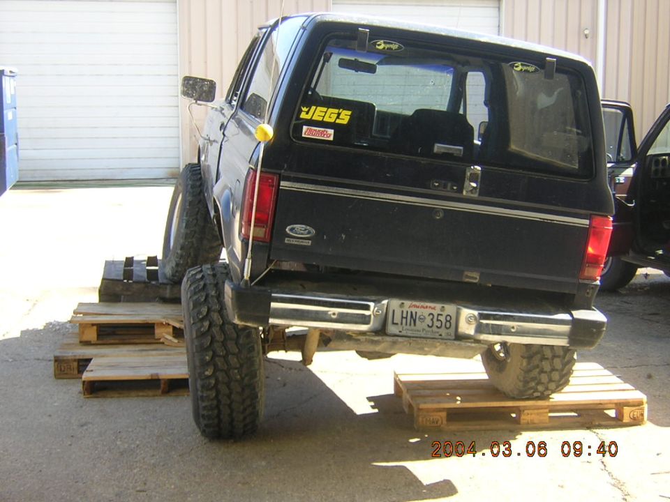 Black Ford Bronco II with lifted suspension, rear view, on wooden pallets. Another vehicle is partially visible to the right