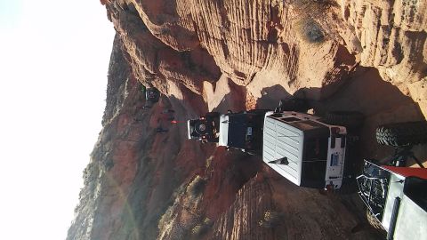 1979 Ford Bronco - Several off-road vehicles are seen traversing a challenging, narrow path along a steep cliff face in a desert landscape.