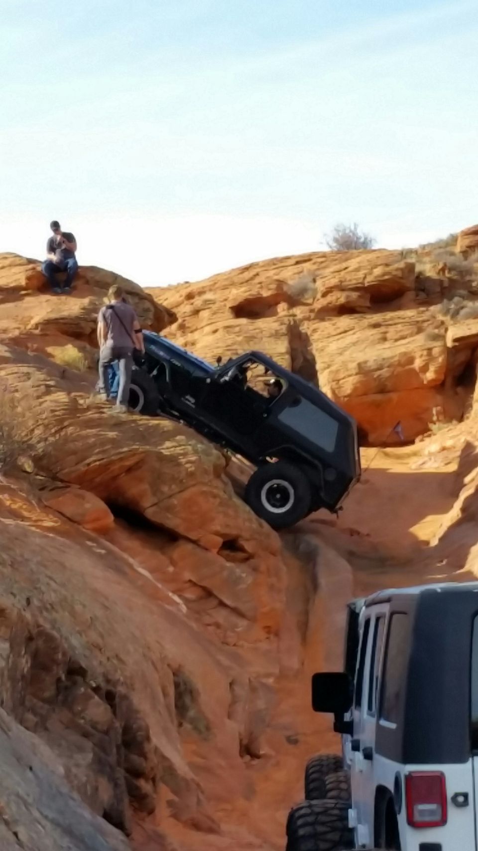 Black Jeep Wrangler attempting to climb a steep, rocky trail with two people watching. Another Jeep is visible in the foreground