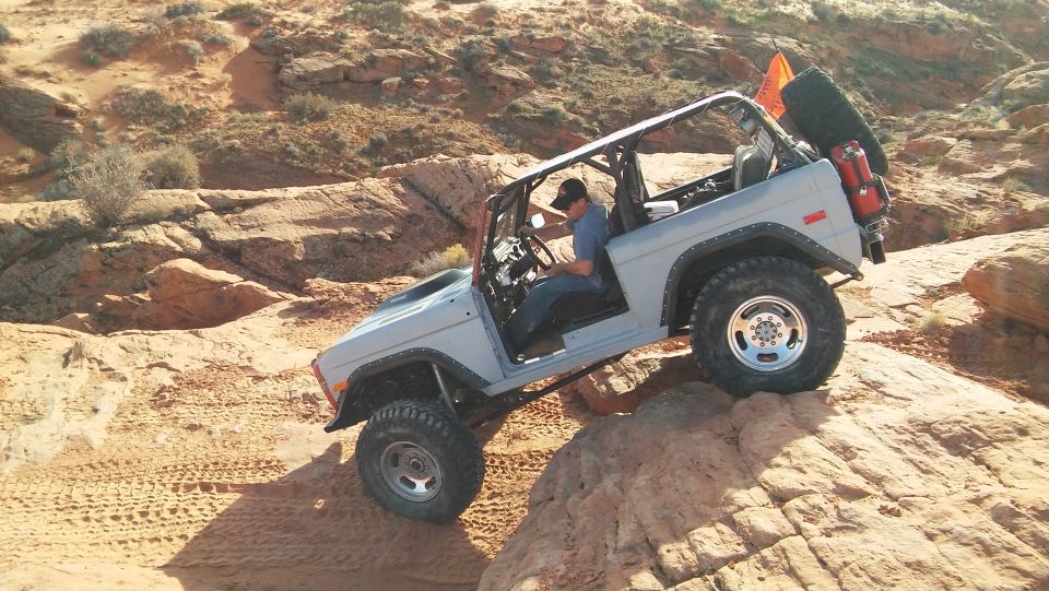 Gray Ford Bronco climbing a rock face, driver visible. Spare tire and gas cans mounted at rear