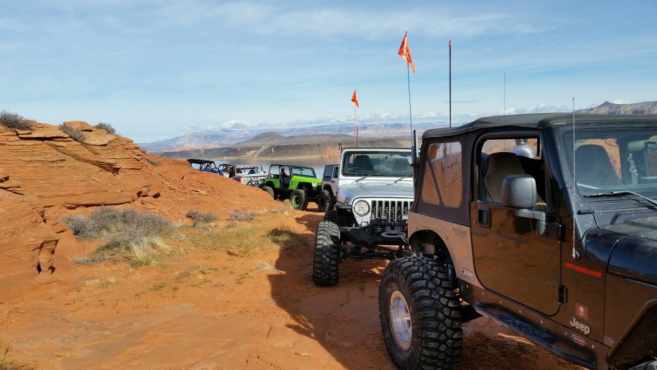 Several Jeeps lined up on a dirt road next to a red rock formation with a lake in the background
