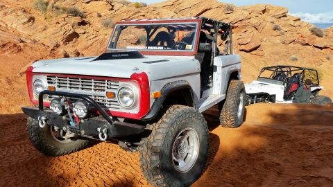 1979 Ford Bronco - Two off-road vehicles, including a modified Ford Bronco, navigate a sandy desert terrain with red rock formations.