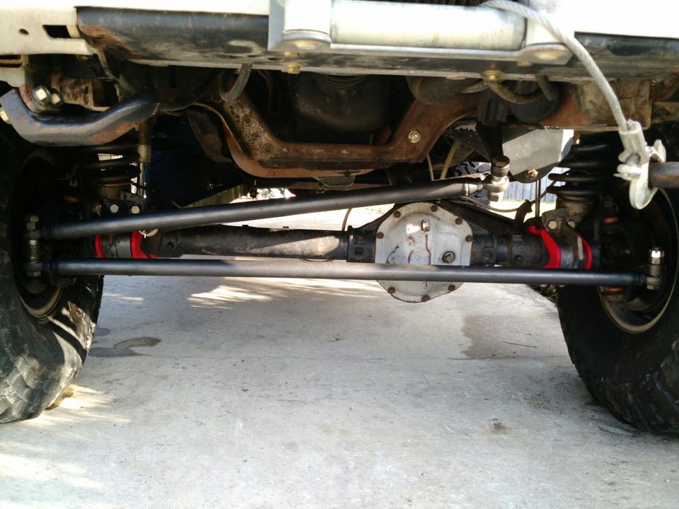 Underside view of a vehicle's front axle, steering components, and suspension. Shows the differential housing and tie rods with red bushings