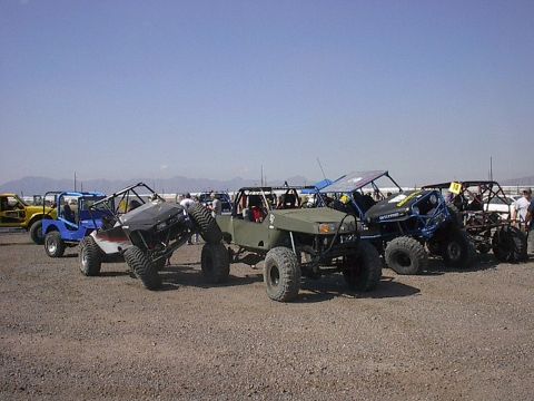 1984 Ford Bronco - A collection of modified off-road vehicles are gathered on a dusty plain with mountains in the distance.