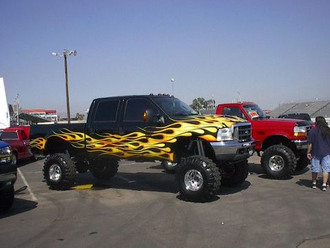 1984 Ford Bronco - A black pickup truck with flame graphics and a lifted suspension is parked next to a red pickup truck.
