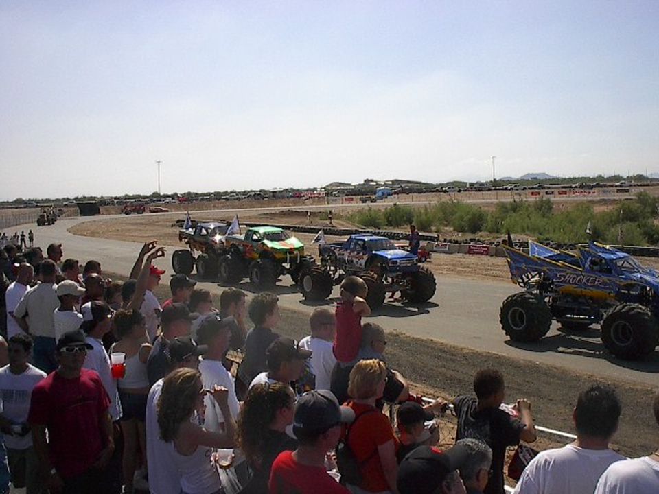Row of monster trucks lined up on dirt track with large crowd watching at outdoor desert event
