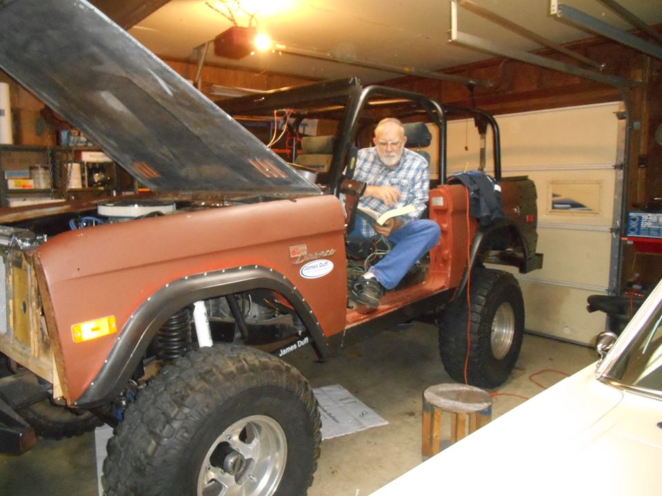 Rusty orange 1972 Ford Bronco in a garage, hood open, with a man reading inside