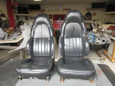 Two black leather car seats sit in a garage with a car body in the background