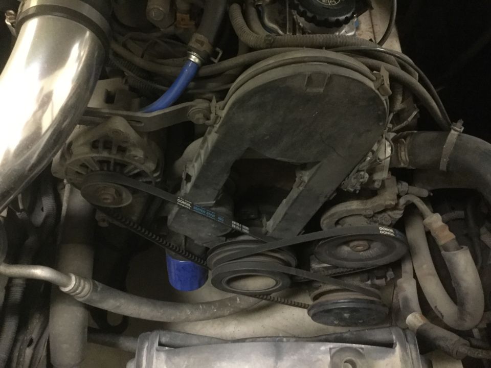 Engine bay with alternator, belts, pulleys, and blue oil filter. Some hoses and wiring are visible