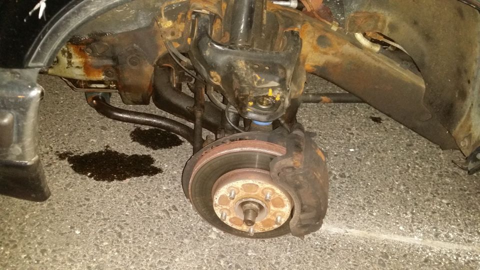 close-up of rusty front suspension and brake components, with fluid leak on pavement