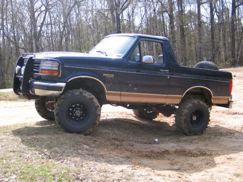1995 Ford Bronco - A lifted Ford Bronco with a brush guard and large tires sits on a dirt road surrounded by trees.
