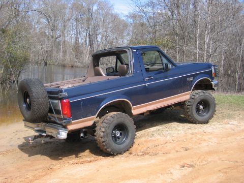 1995 Ford Bronco - This lifted Ford Bronco is parked on a dirt path next to a body of water with trees in the background.