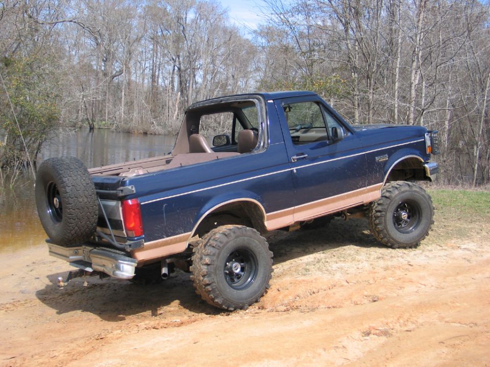 Blue Ford Bronco with lifted suspension and spare tire on the back, parked near water