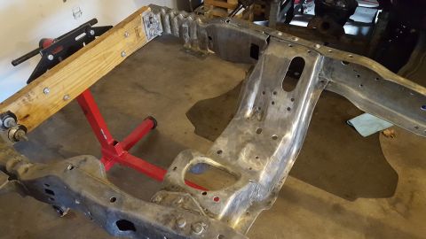 1994 Ford Bronco - A bare metal, stripped-down vehicle frame is supported by a red engine stand and a wooden beam. The frame appears to be undergoing restorat…