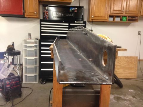 1979 Ford Bronco - A large, welded metal channel sits on a wooden workbench in a garage. The channel is angled and has a circular cutout on its side. Various…