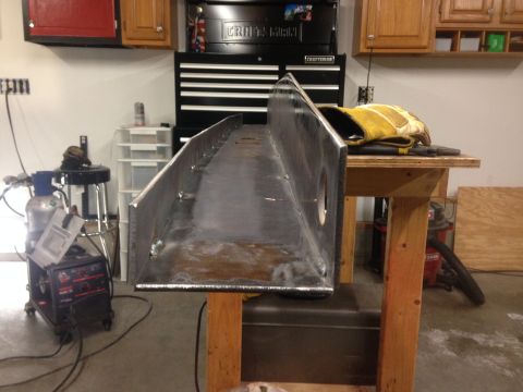 1979 Ford Bronco - A large, welded metal beam is positioned on a wooden workbench in a garage setting. A welding machine, gas cylinder, and storage cabinets a…