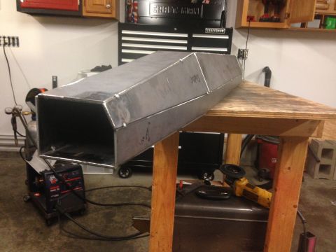 1979 Ford Bronco - A custom-fabricated metal duct or enclosure is shown resting on a wooden workbench, with welding equipment and tools visible in the backgro…
