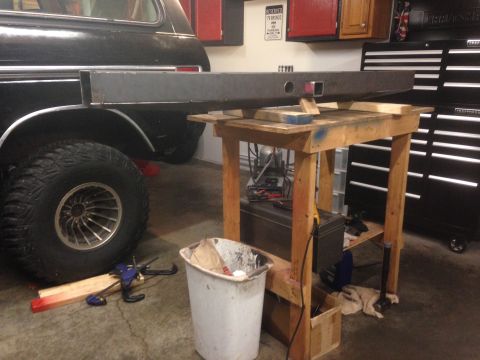 1979 Ford Bronco - A custom steel bumper is supported by a wooden workbench next to the rear fender and tire of a black SUV. The bumper appears to be in the p…