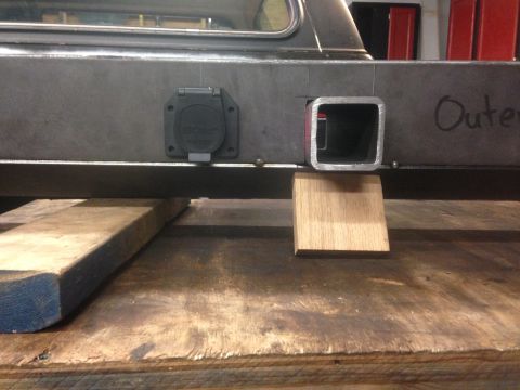 1979 Ford Bronco - A close-up view of the rear of a vehicle frame showing a trailer hitch receiver and a 7-pin trailer connector. A small wooden block is supp…