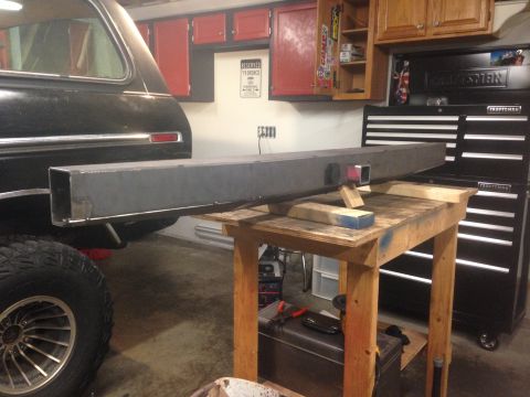 1979 Ford Bronco - A custom-fabricated steel bumper is shown resting on a wooden workbench in a garage setting, with the rear of a black SUV visible to the le…
