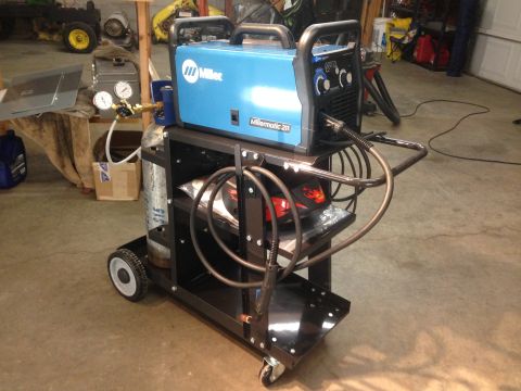 1979 Ford Bronco - A blue Miller welding machine is on a black metal cart with a gas cylinder. The cart has wheels and shelves holding welding equipment.