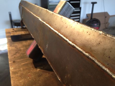 1979 Ford Bronco - A close-up shot of a welded metal beam resting on a wooden surface. The beam shows signs of rust and has visible weld lines.