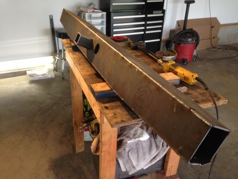 1979 Ford Bronco - A long, heavy steel beam with cutouts and a circled number '2' drawn on it rests on a wooden workbench. A yellow angle grinder and a red sh…