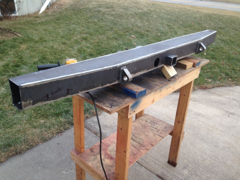 1979 Ford Bronco - A custom fabricated steel bumper is resting on a wooden workbench outdoors. The bumper has several welded brackets and holes cut into it, a…