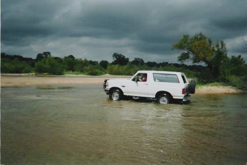 white 1995 Ford Bronco driving through a river with a spare tire on the back