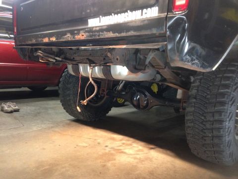 1979 Ford Bronco - The rear underside of a black pickup truck is shown, with a large fuel tank visible. A pair of athletic shoes rests on the concrete floor t…