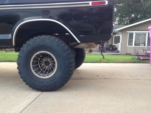1979 Ford Bronco - The rear driver's side of a black lifted Ford Bronco is shown parked on a concrete driveway. The vehicle has large off-road tires and custo…