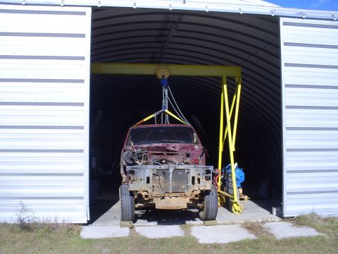 1992 Ford Bronco - The front end of a maroon SUV is suspended by a yellow gantry crane inside a large metal building.