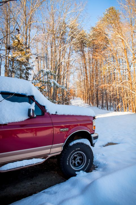 1995 Ford Bronco - A red Ford Bronco sits partially covered in snow on a bright, sunny day with trees in the background.