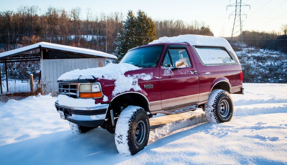 Red 1995 Ford Bronco covered in snow, parked in a snowy area with a small building and trees in the background