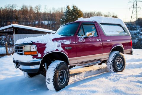 1995 Ford Bronco - A maroon Ford Bronco with a tan stripe is partially covered in snow, parked in a winter landscape.