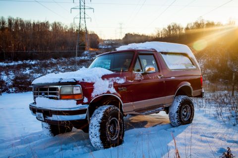 1995 Ford Bronco - A maroon Ford Bronco sits covered in snow during a winter day, with power lines in the background.