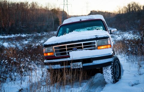 1995 Ford Bronco - A red Ford truck is partially covered in snow, driving through a snowy field with trees in the background.