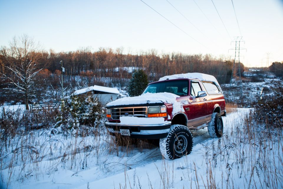 Red 1995 Ford Bronco parked in a snowy field with trees and a small building in the background