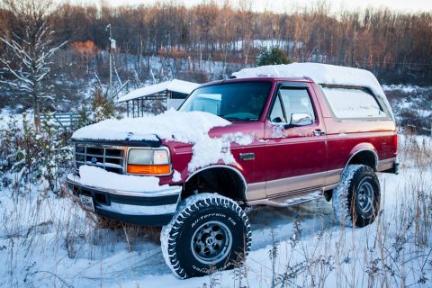 1995 Ford Bronco - A red Ford Bronco with tan trim sits in a snowy field, its tires and hood covered in snow.