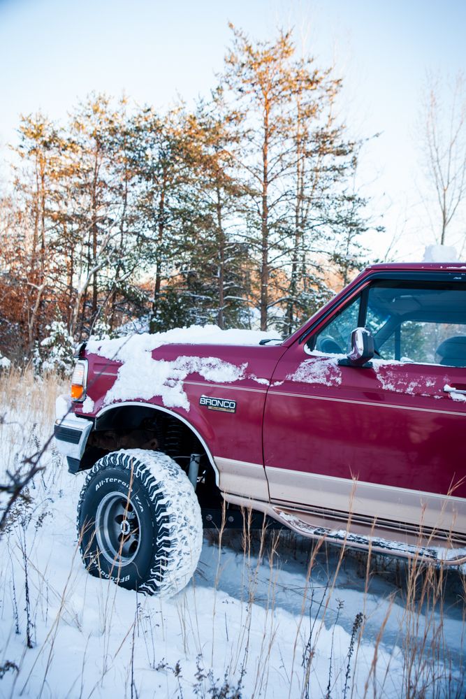 Red 1995 Ford Bronco with snow on hood and fender, driving in a snowy field