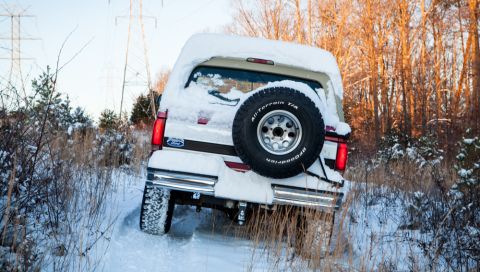 1995 Ford Bronco - A Ford Bronco navigates a snow-covered trail surrounded by trees and power lines.