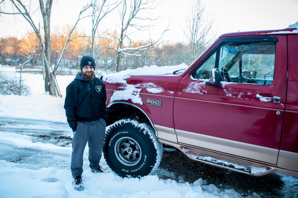 Man in Raiders gear stands by a red 1995 Ford Bronco covered in snow